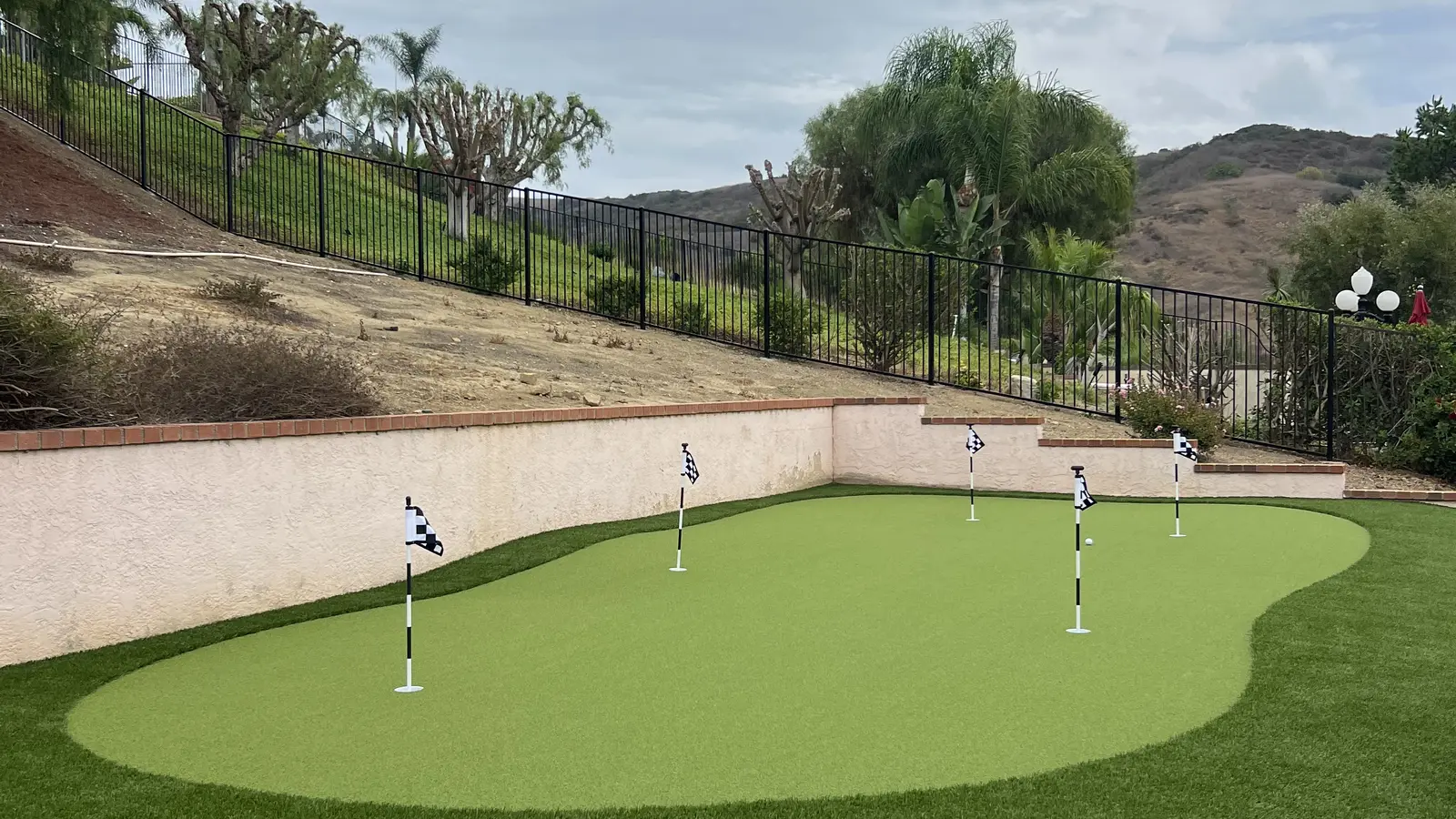 Backyard putting green with five flags below a fenced hillside